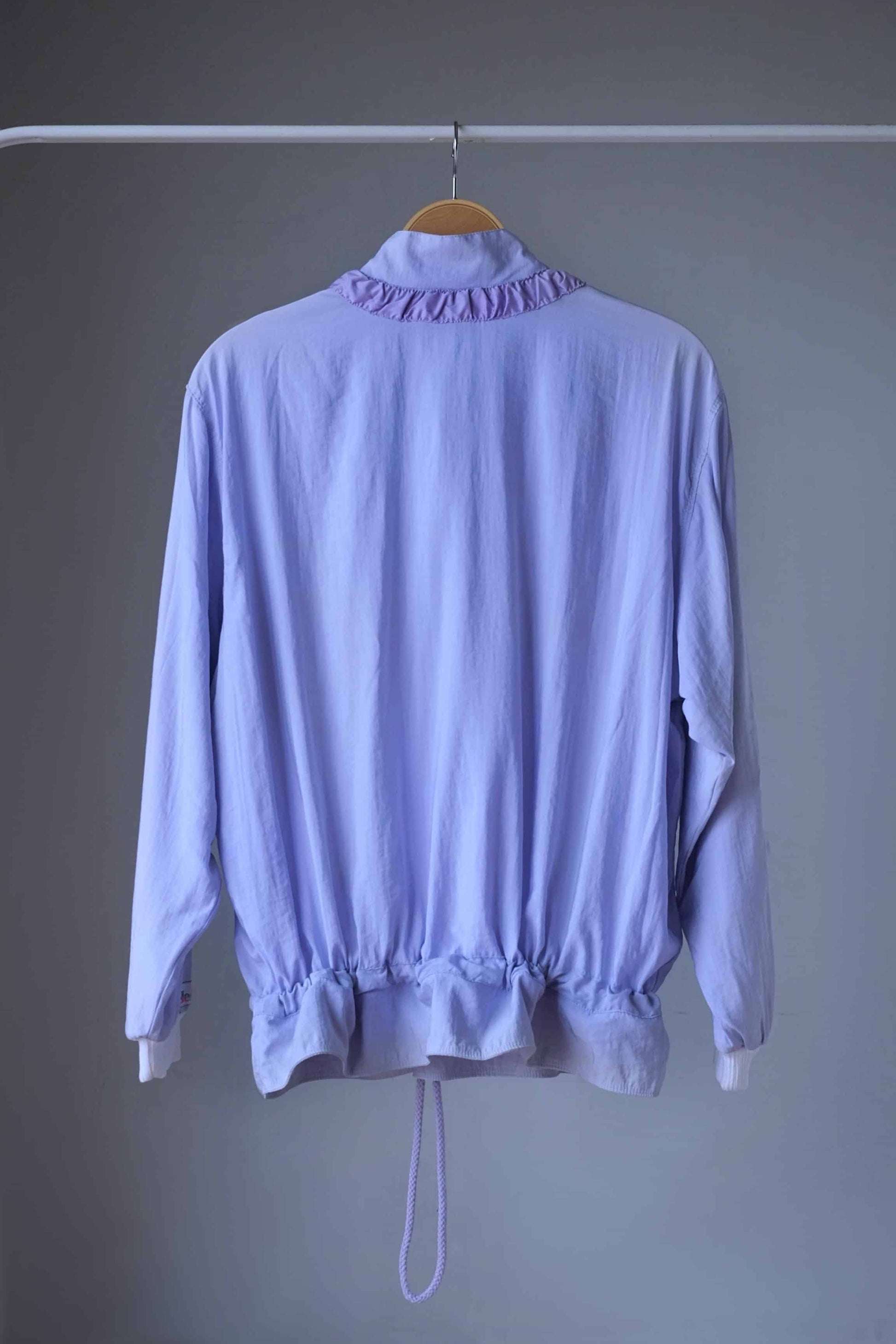 Vintage Ellesse jacket in pastel purple with a drawstring hem, seen from the back and hanging on a hanger.