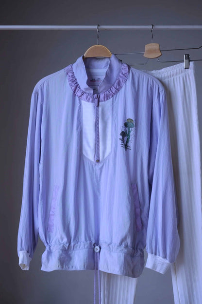 Close up of a Vintage Ellesse tracksuit in pastel purple and white, consisting of a cropped windbreaker with floral print and ribbed white leggings, hanging on a hanger with metal clips.