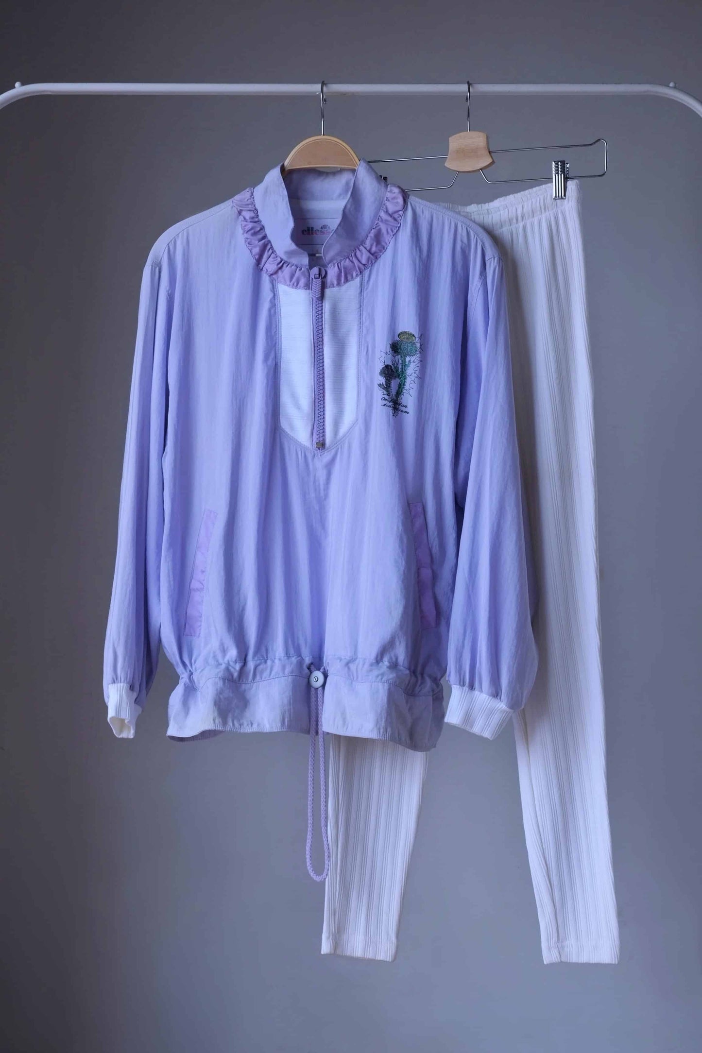 Vintage Ellesse tracksuit in pastel purple and white, consisting of a cropped windbreaker with floral print and ribbed white leggings, hanging on a hanger with metal clips.