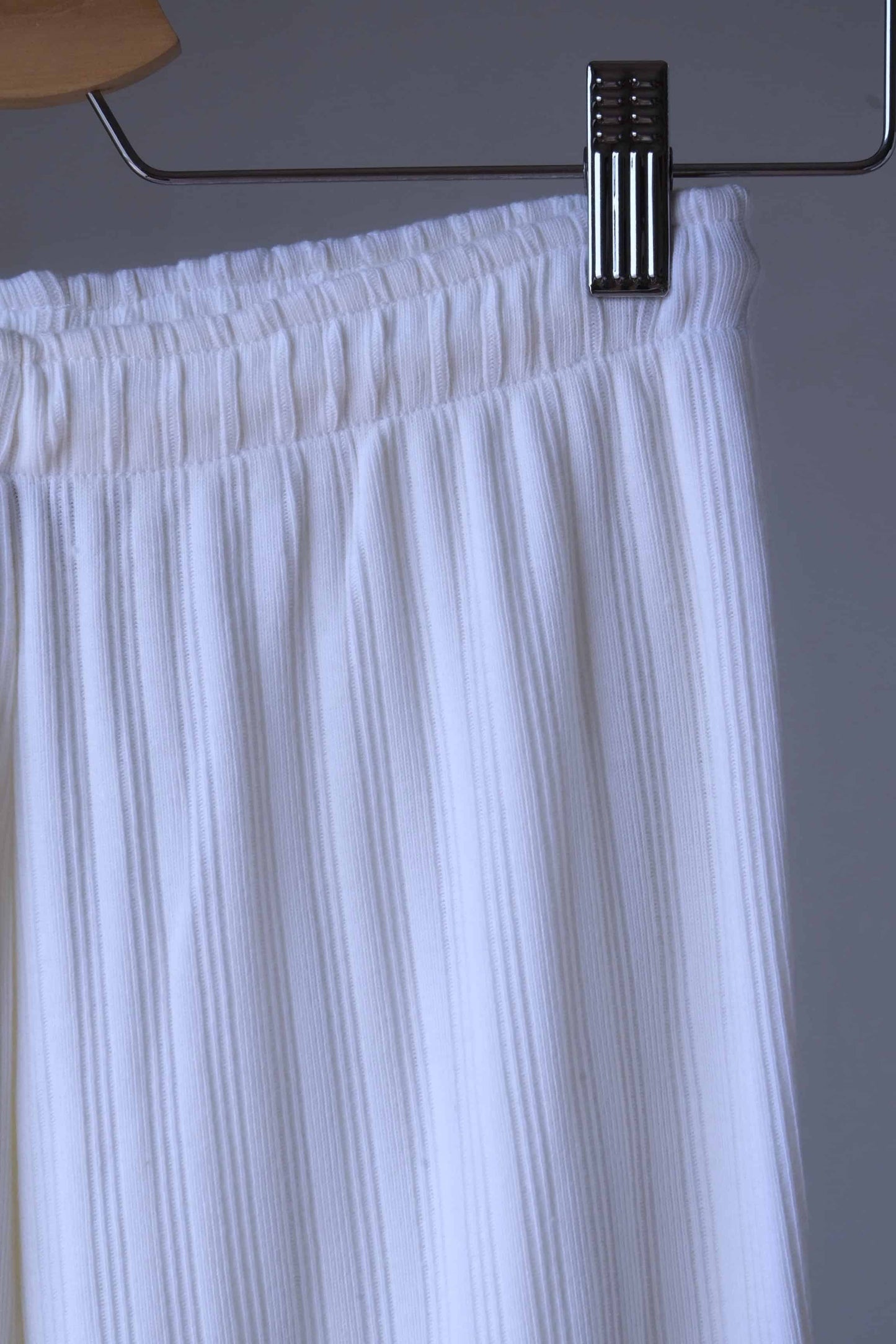 Close-up of white ribbed leggings with an elastic waistband, hanging on a hanger with metal clips.