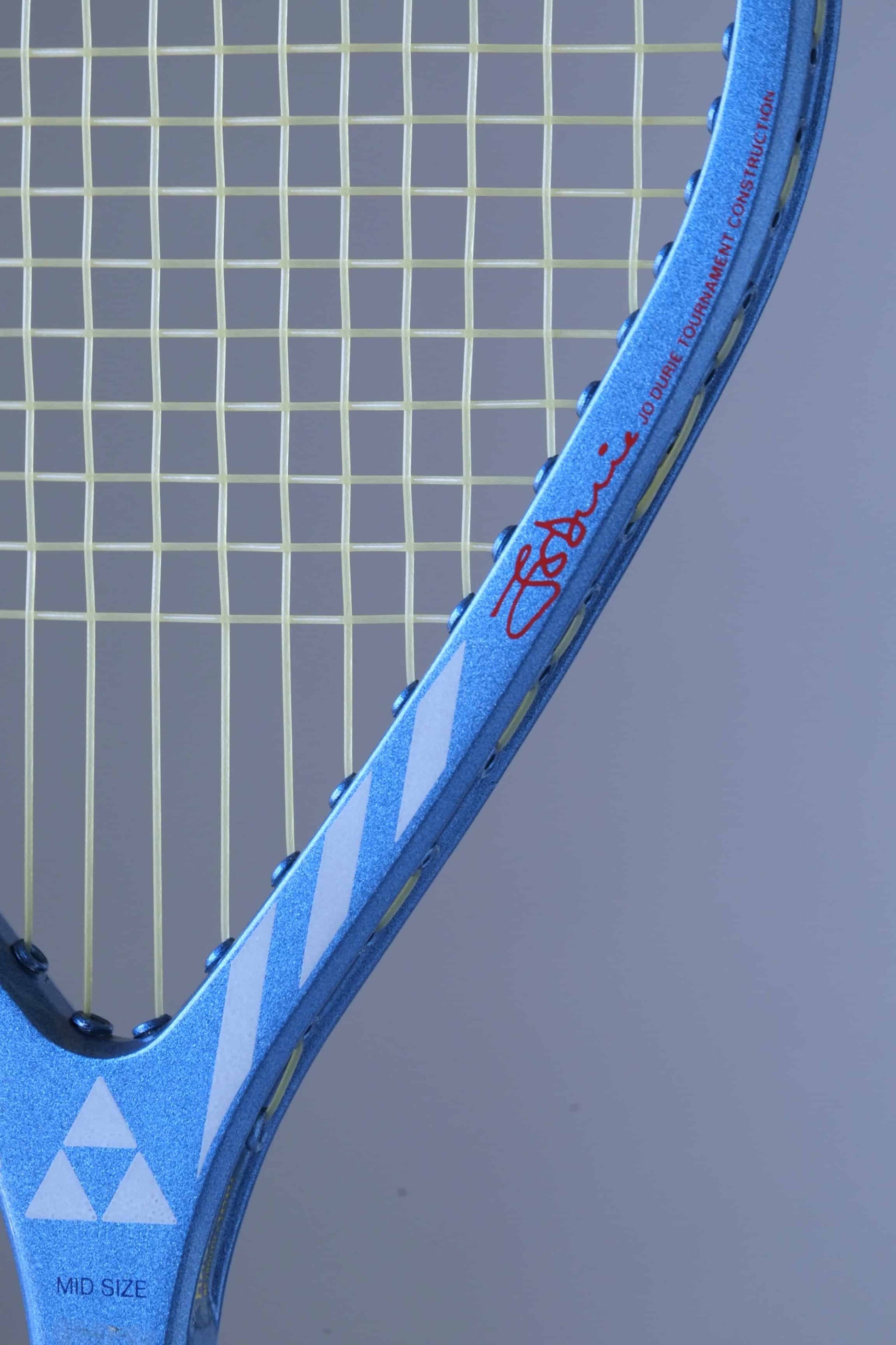 Close-up of a Fischer tennis racket handle with a blue and white diamond pattern.
 The word "MID SIZE" is written in white on the handle. The signature "JO DURIE TOURNAMENT CONSTRUCTION" is also visible in white.