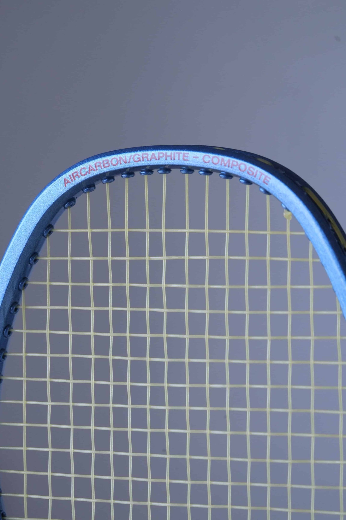 Close-up of a Fischer tennis racket head with yellow strings.
 The frame is blue and has the text "AIRCARBON/GRAPHITE-COMPOSITE" written in red.