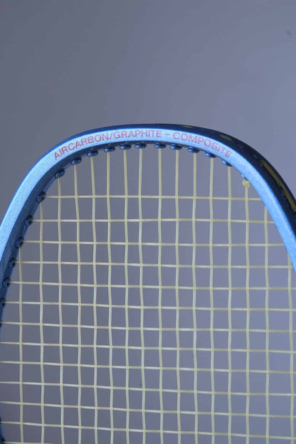 Close-up of a Fischer tennis racket head with yellow strings.
 The frame is blue and has the text "AIRCARBON/GRAPHITE-COMPOSITE" written in red.