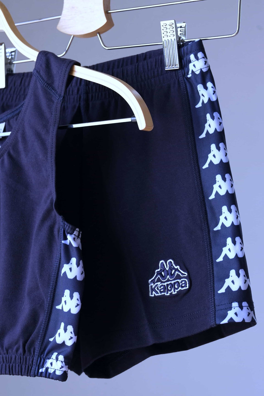 Close-up view of a navy blue KAPPA vintage women's athletic crop top and shorts set on hangers, highlighting the white KAPPA logo taping on the sides of both garments and the KAPPA logo on the shorts.