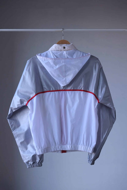 Back view of a vintage Koflach windbreaker jacket in white and silver with a foldable hood unfolded, showcasing its red trim detail and elastic hem, hung on a wooden hanger.