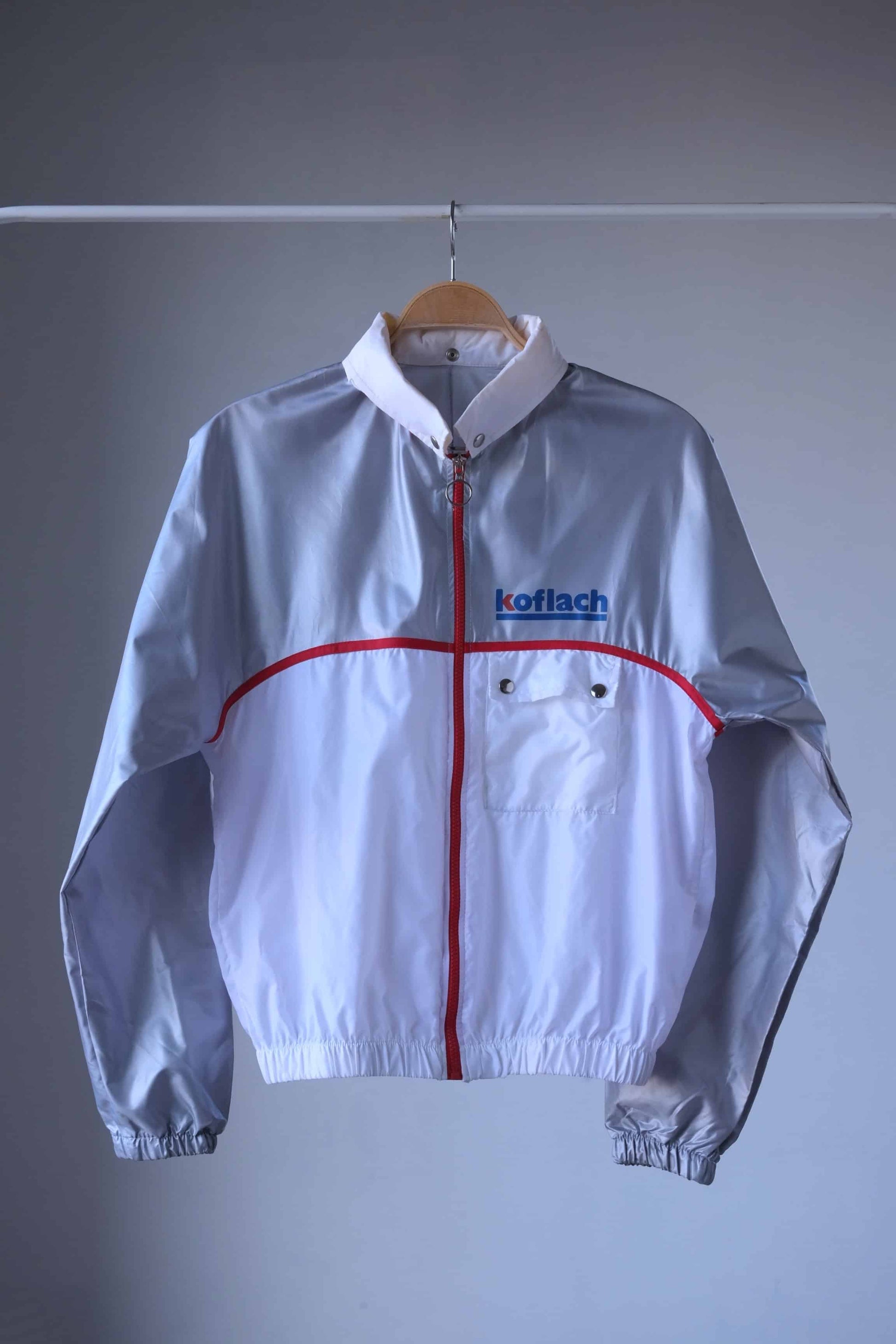 Front view of a vintage Koflach windbreaker jacket in white and silver with red accents, featuring a zippered closure, snap pocket, and elastic cuffs, displayed on a wooden hanger.