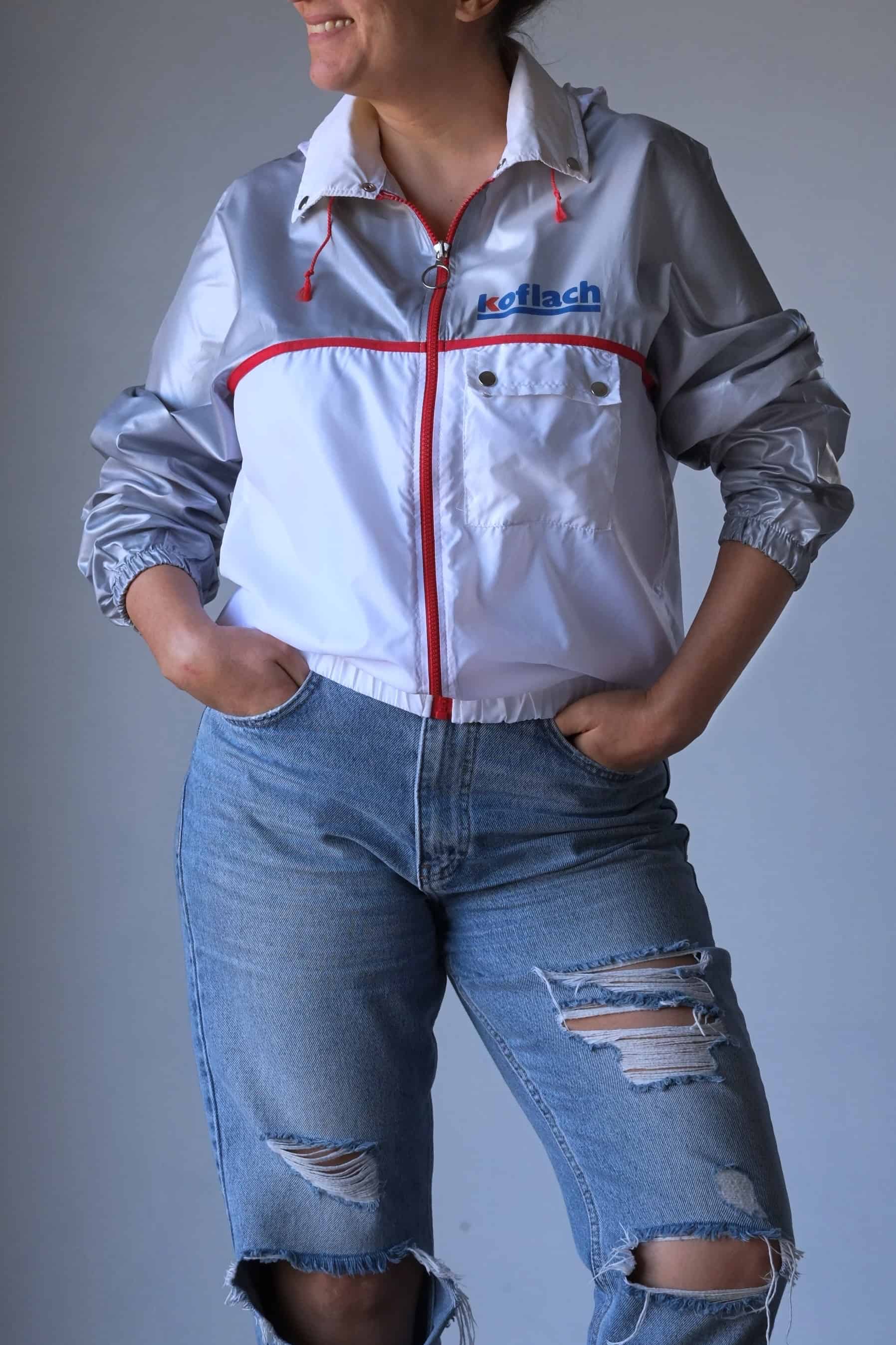 A model wearing a vintage Koflach windbreaker jacket featuring a white and silver color scheme with red accents. The jacket has a foldable hood tucked into the collar, a front pocket with snap closures, and elastic cuffs and hem. The model pairs the jacket with distressed denim shorts and is smiling, with hands casually in her pockets.