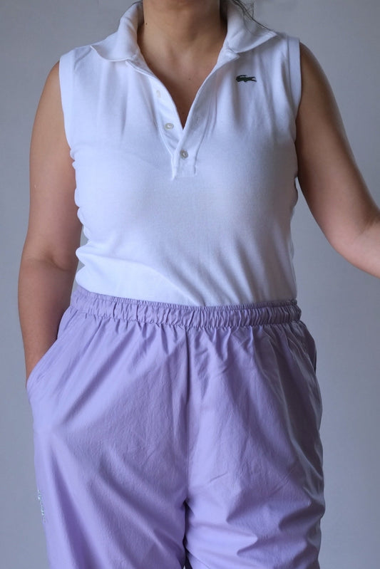 A woman wearing a vintage white sleeveless Lacoste polo shirt, showcasing the shirt's fit and style. The shirt features the signature green Lacoste crocodile logo on the chest. She is also wearing light purple elastic-waist pants, standing against a neutral background.