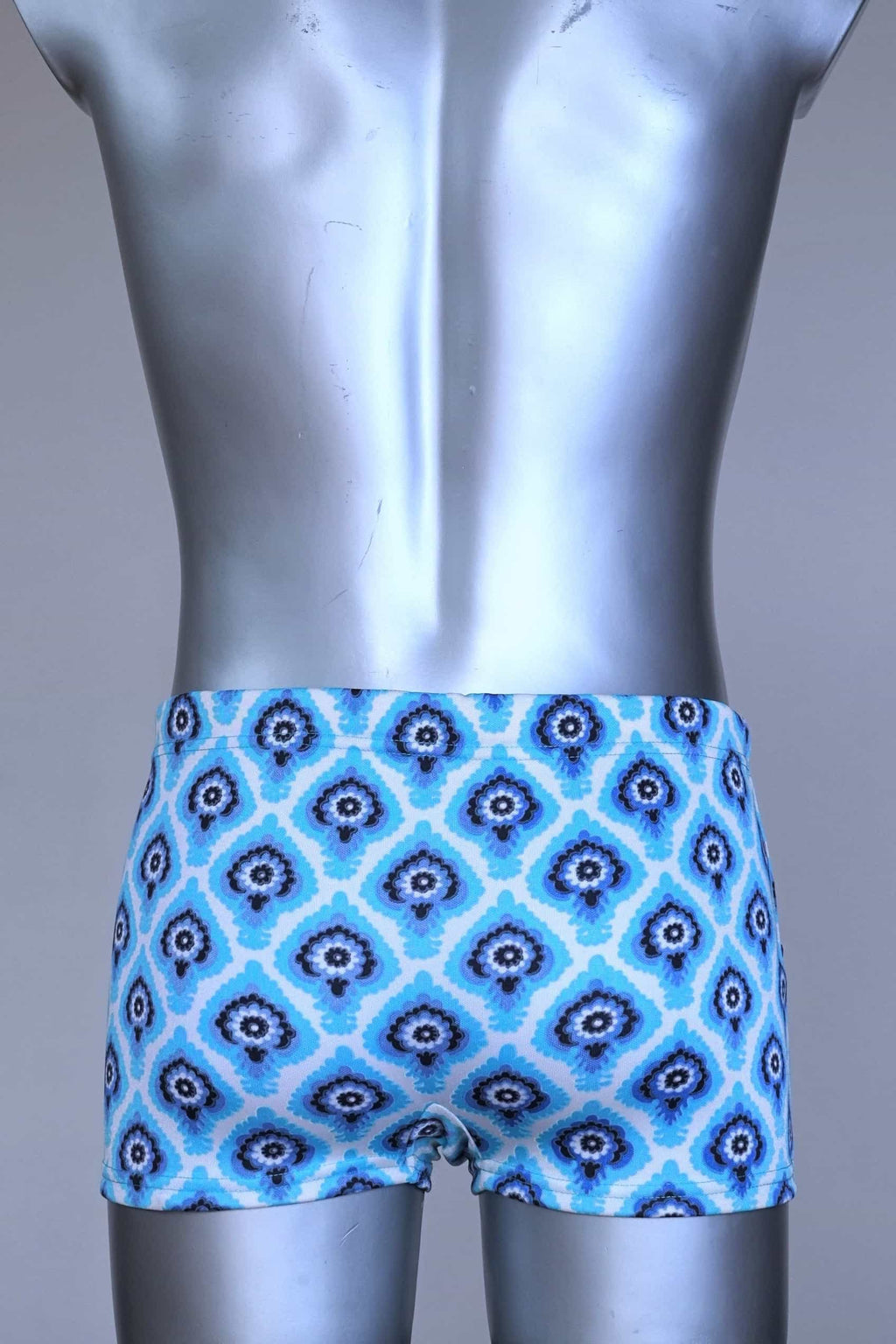 Back view of vintage 60s Lahco swim trunks in icy blue with a repeating dark blue and white geometric medallion pattern, featuring a zip pocket and brand tag.