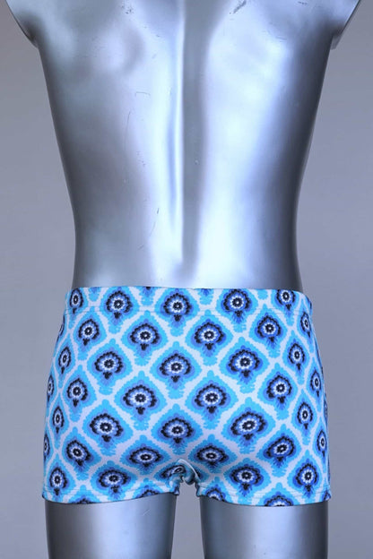 Back view of vintage 60s Lahco swim trunks in icy blue with a repeating dark blue and white geometric medallion pattern, featuring a zip pocket and brand tag.