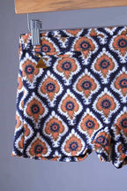 Close view of a Vintage 60s Lahco swim trunks in brown with a geometric medallion print, displayed on a wooden hanger, featuring a front drawstring and zip pocket.