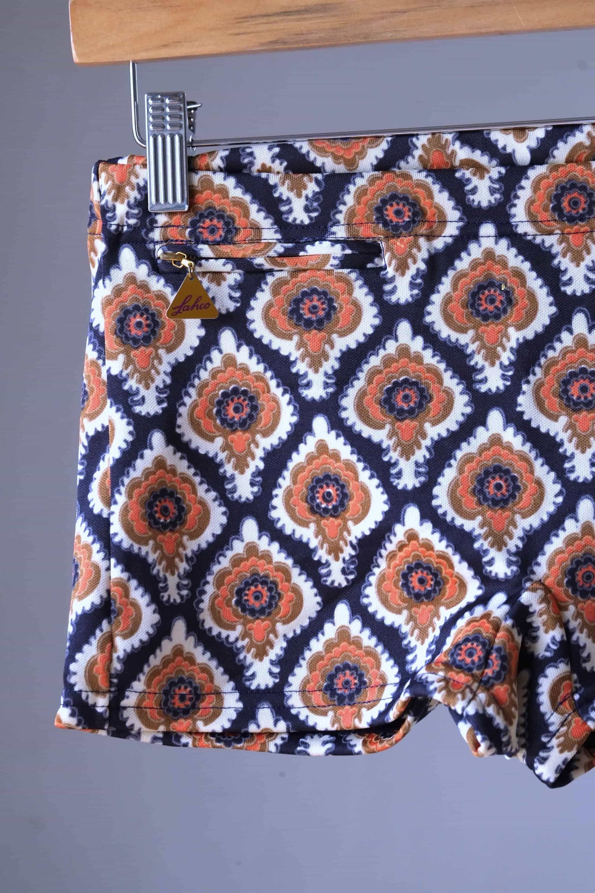 Close view of a Vintage 60s Lahco swim trunks in brown with a geometric medallion print, displayed on a wooden hanger, featuring a front drawstring and zip pocket.