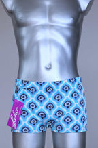 Front view of vintage 60s Lahco swim trunks in icy blue with a repeating dark blue and white geometric medallion pattern, featuring a zip pocket and brand tag.