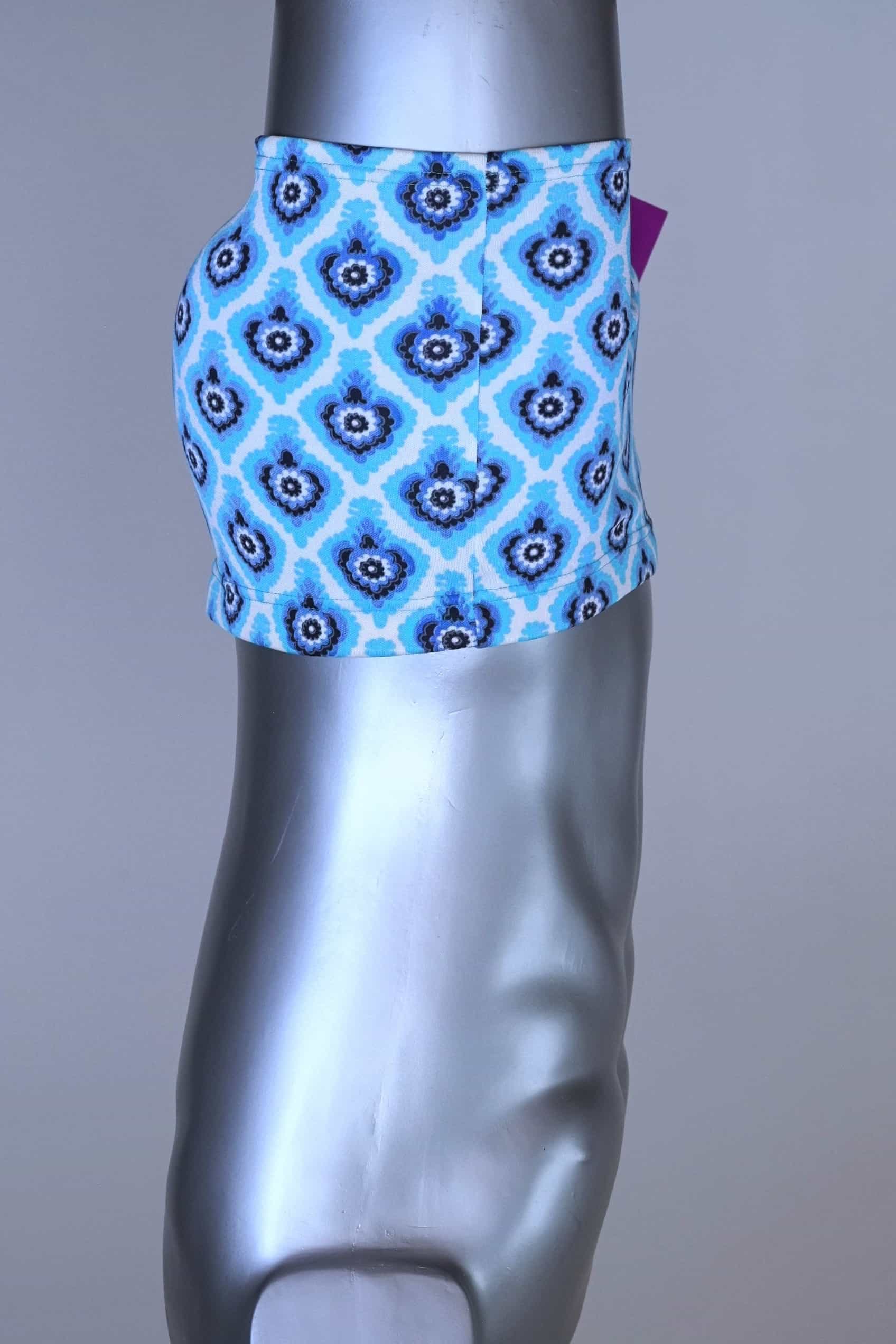 Side view of vintage 60s Lahco swim trunks in icy blue with a repeating dark blue and white geometric medallion pattern, featuring a zip pocket and brand tag.
