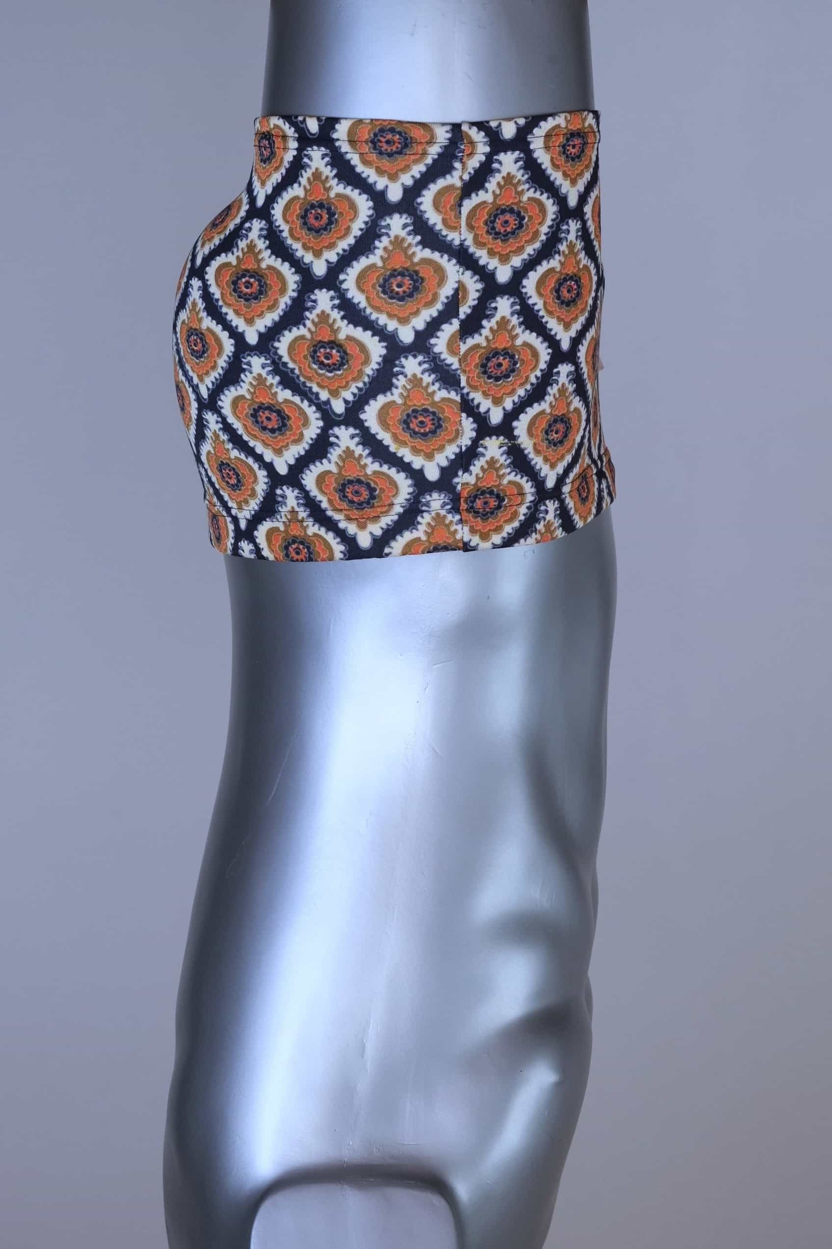 Side view of vintage 60s Lahco swim trunks in navy with an allover brown, orange, and cream geometric medallion print, featuring a zip pocket and original brand tag.