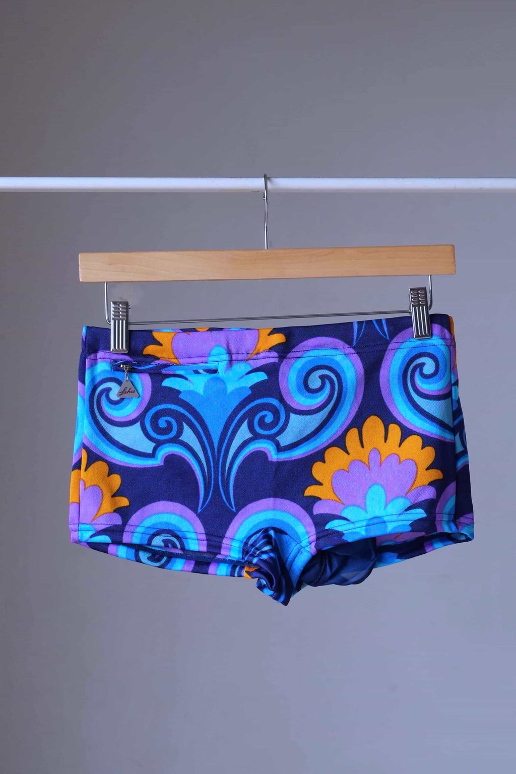 LAHCO vintage 60s square-cut swim trunks in blue, purple, and orange psychedelic print, hanging on a wooden hanger.