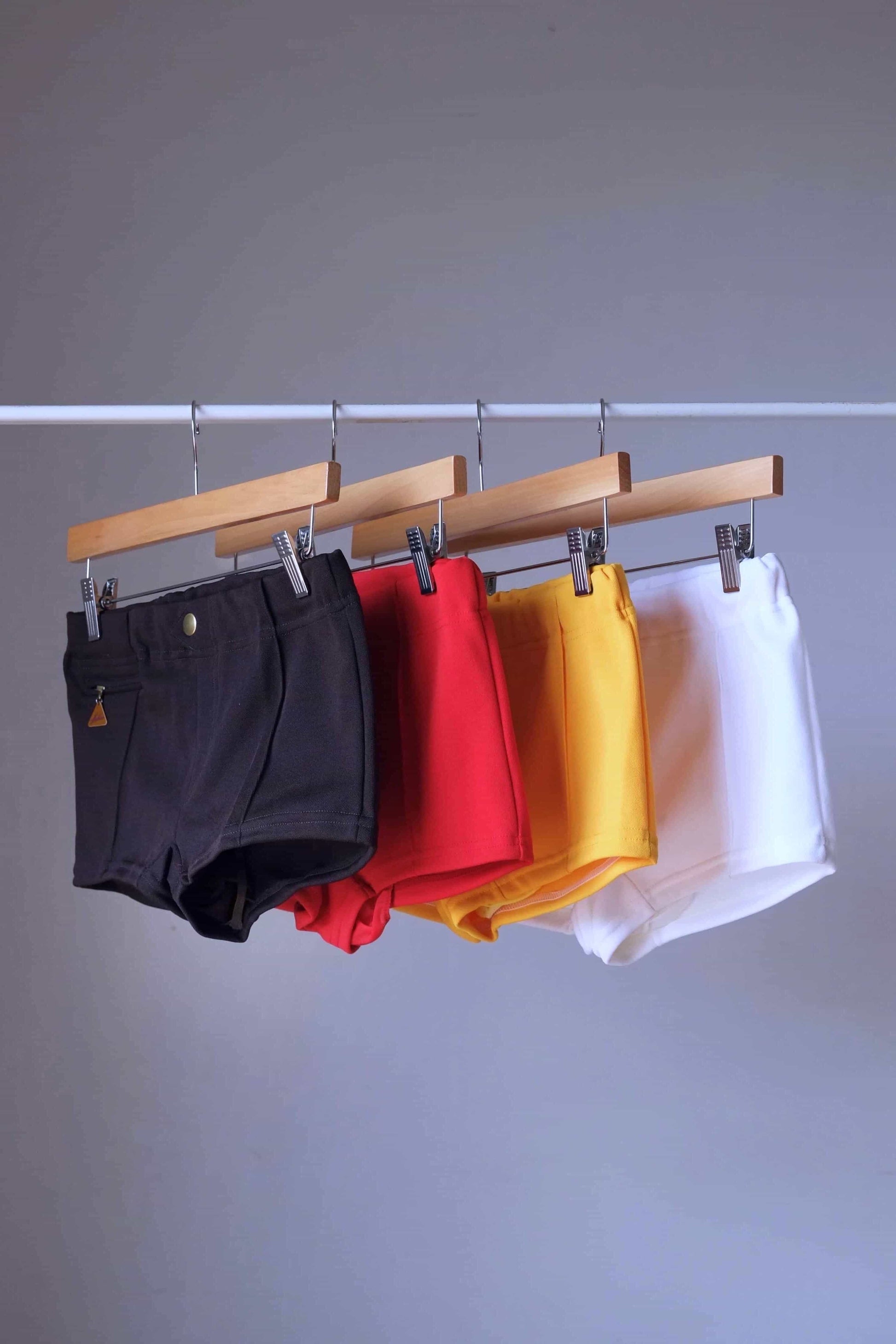 Four color options of Lahco Mataro vintage 60s swim trunks (brown, red, yellow, white) hanging on wooden hangers.