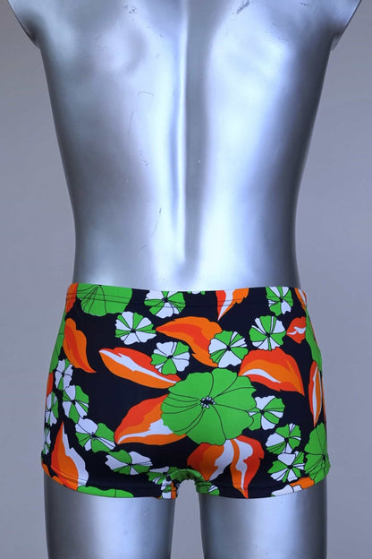 Back view of LAHCO vintage 1960s men's swim trunks on a silver mannequin. The trunks are black with a vibrant green, orange, and white floral pattern.
