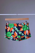 Front view of LAHCO vintage 1960s men's swim trunks on a wooden hanger. The trunks are black with a vibrant green, orange, and white floral pattern.

