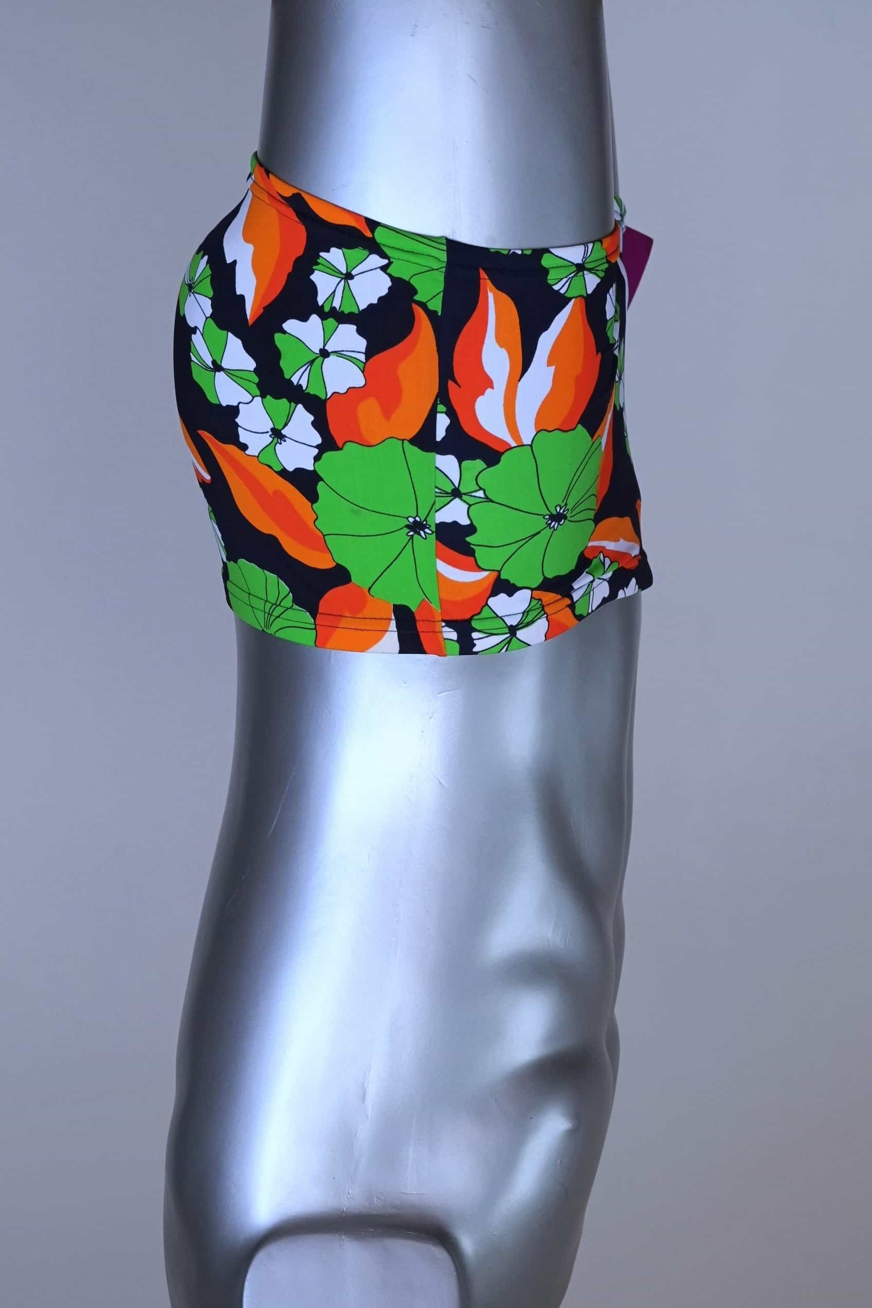 Side view of LAHCO vintage 1960s men's swim trunks on a silver mannequin. The trunks are black with a vibrant green, orange, and white floral pattern.
