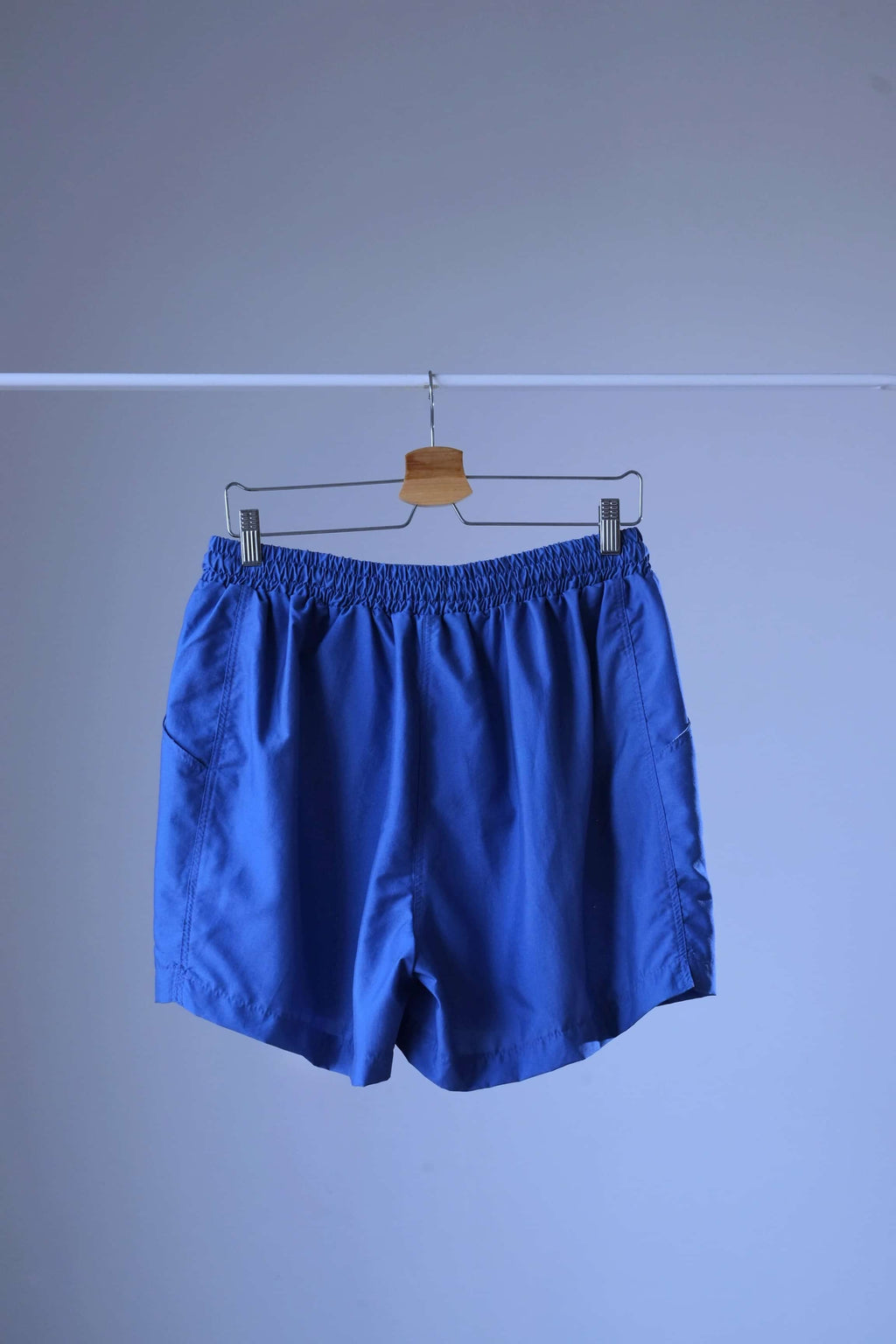 Rear view of a pair of blue vintage Löffler shorts with a small logo on the left leg, hanging on a hanger