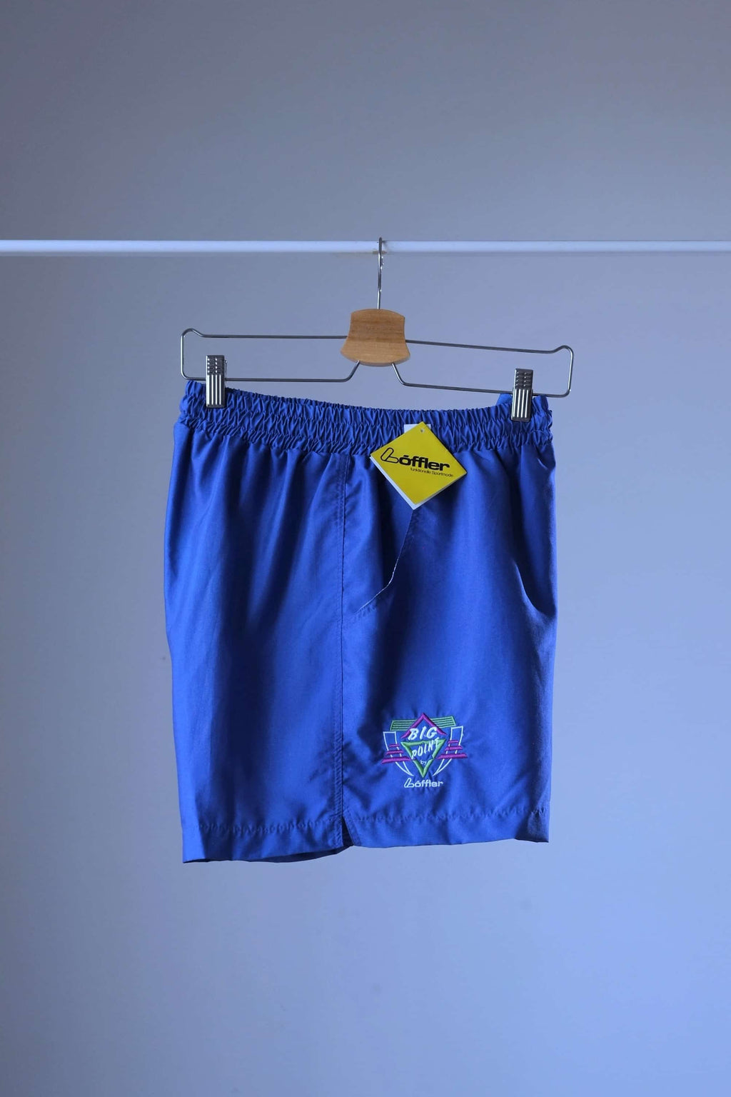 Side view of a pair of blue vintage Löffler shorts with a small logo on the left leg, hanging on a hanger, a yellow Löffler hang tag is visible.
