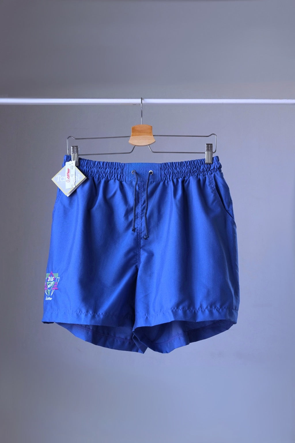 Front view of a pair of blue vintage Löffler shorts with a small logo on the left leg, hanging on a hanger.