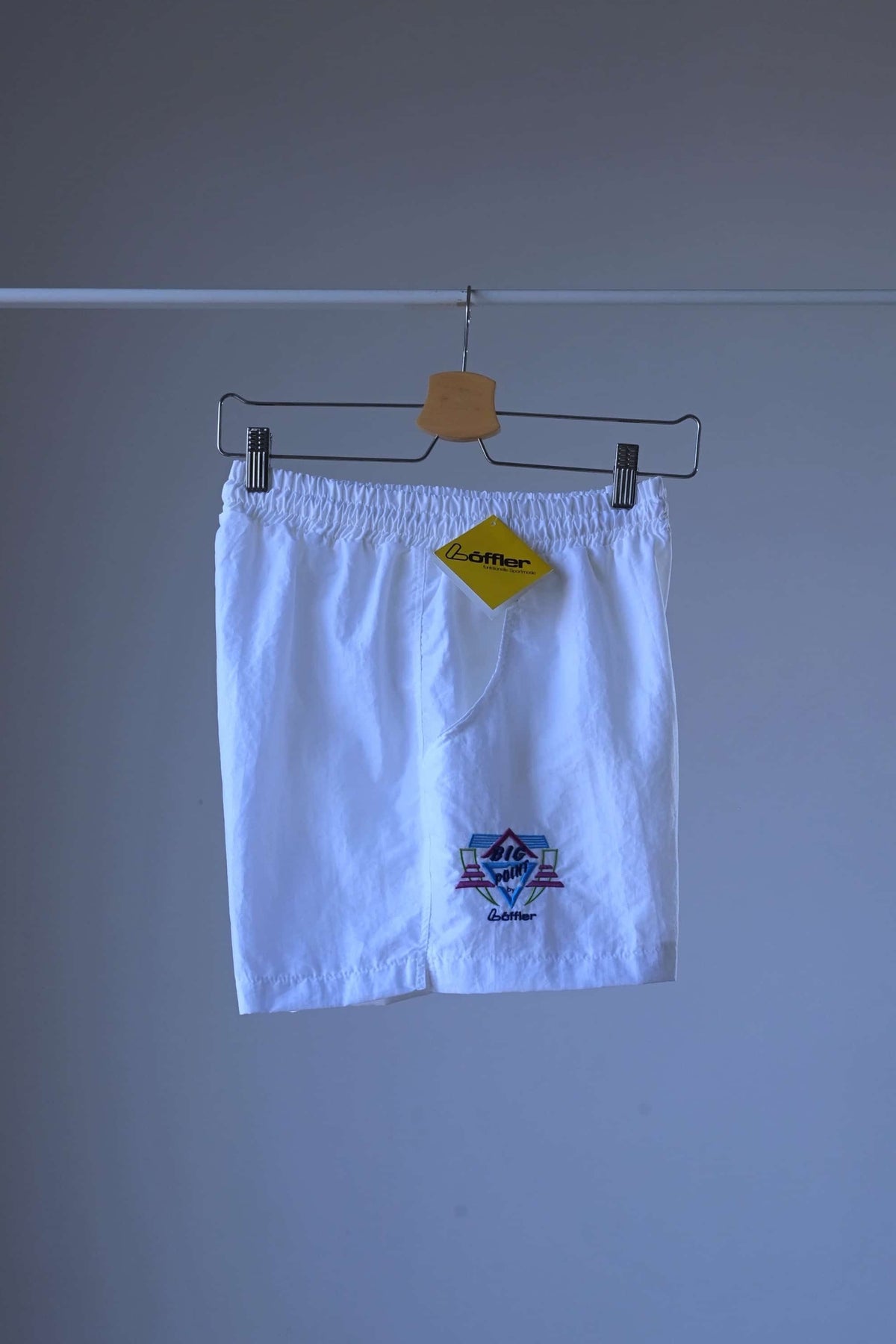Side view of a pair of white vintage Löffler shorts with a small logo on the left leg, hanging on a hanger, a yellow Löffler hang tag is visible.