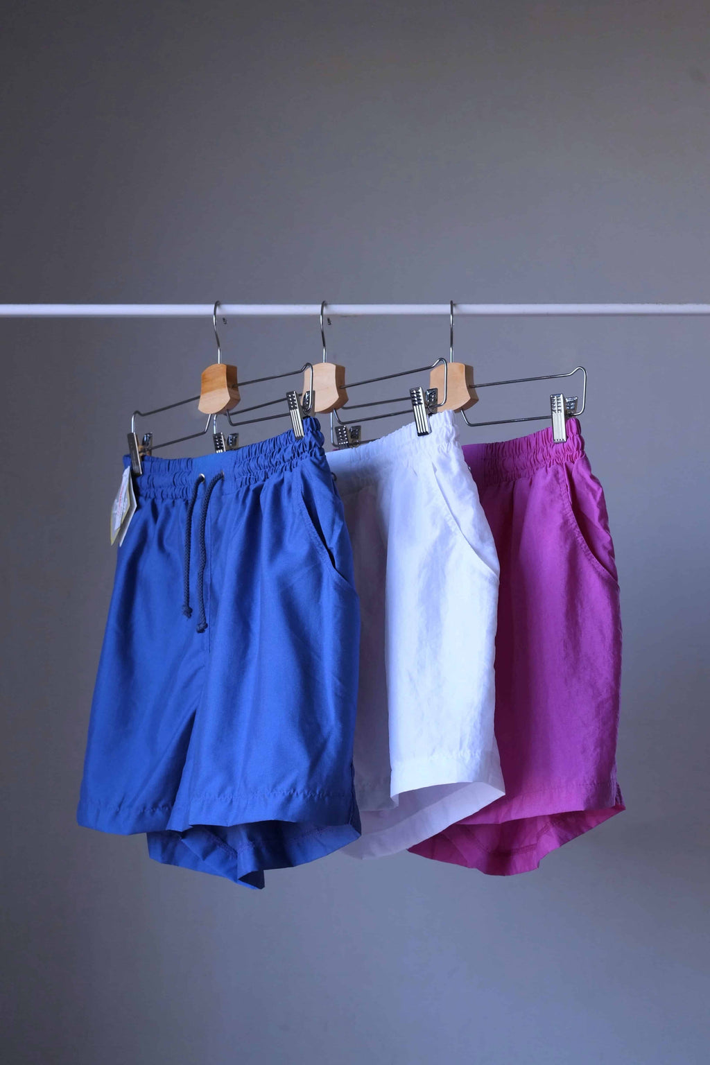 Three pairs of vintage Löffler shorts in blue, white, and magenta hanging together on a clothing rack.