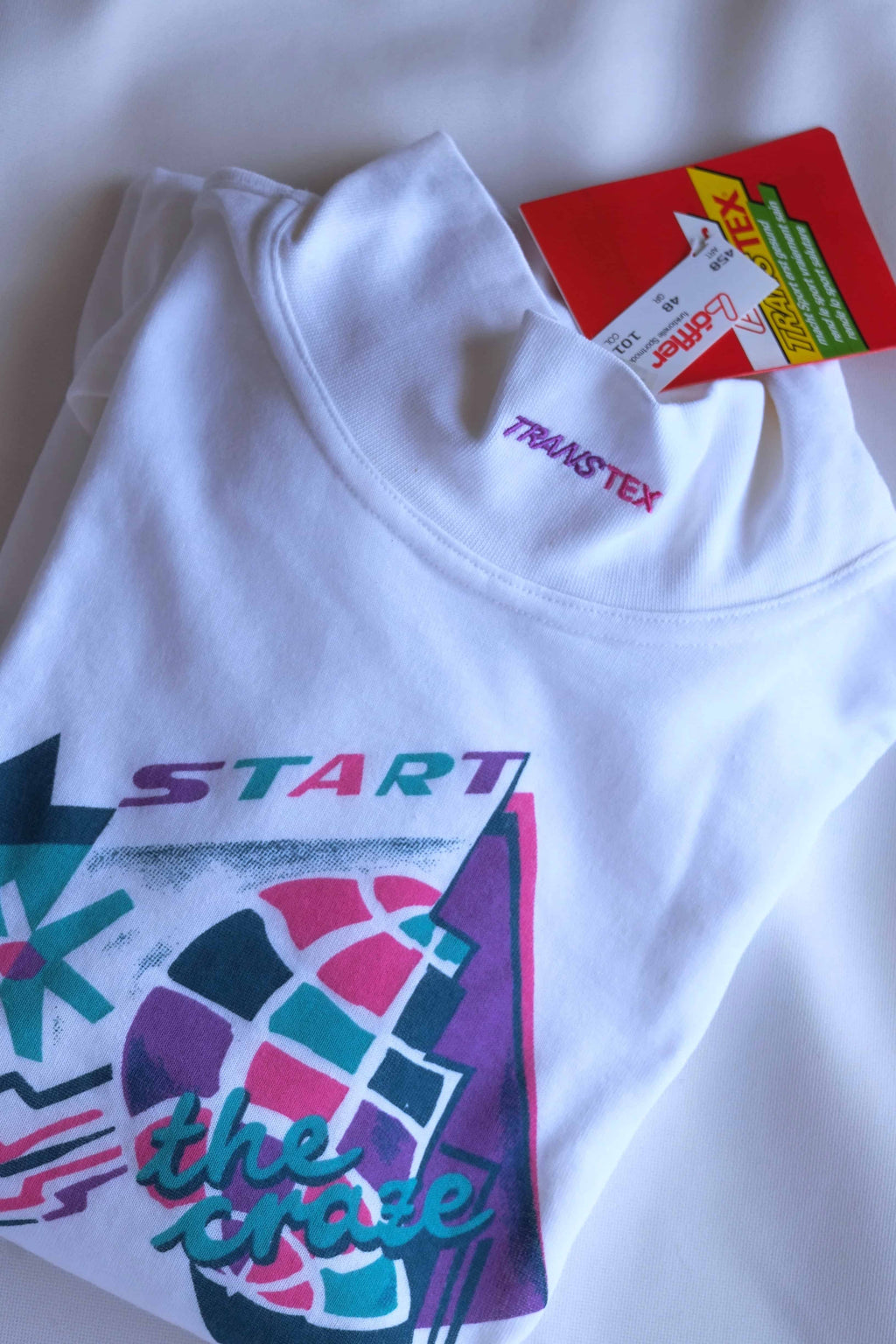 Flat lay of the white Löffler mock neck top showing the pink Transtex collar embroidery, the “Start the Craze” graphic and the red Löffler Transtex hang tag