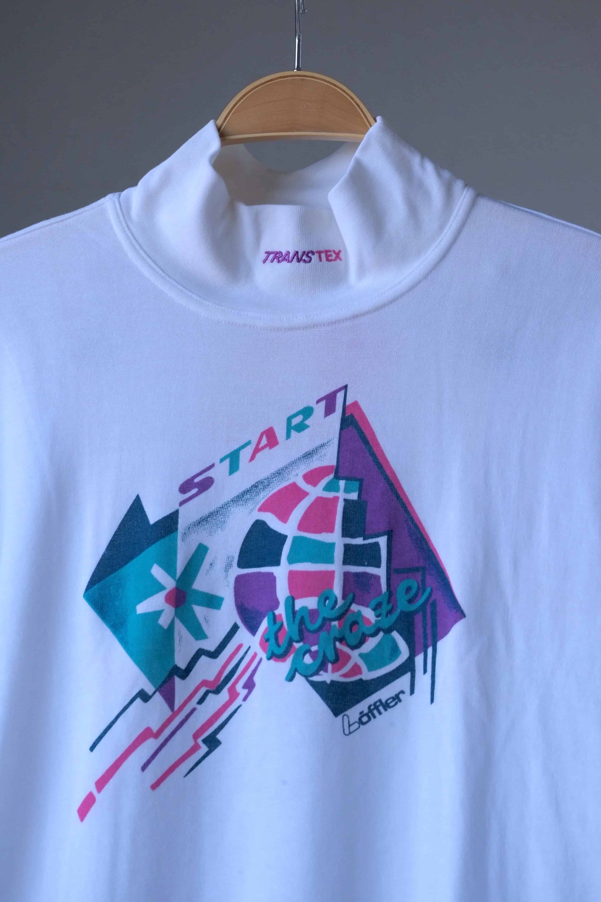 Close-up of the mock neck collar with purple Transtex embroidery above the pink, teal and purple “Start the Craze” graphic on a white Löffler 90s top
