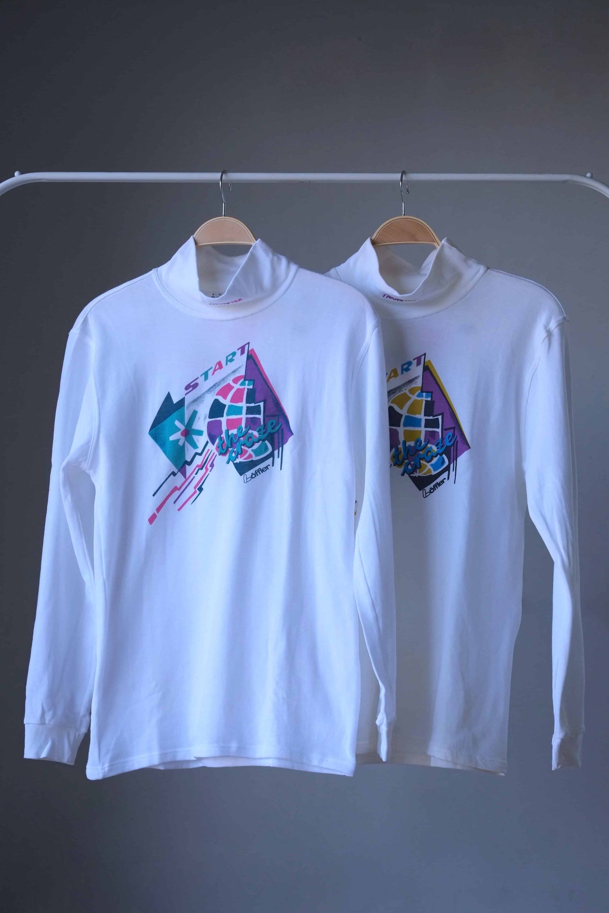 Two white Löffler 90s mock neck long-sleeve tennis tops on hangers, one with a pink, teal and purple “Start the Craze” graphic and one with a yellow, blue and purple version