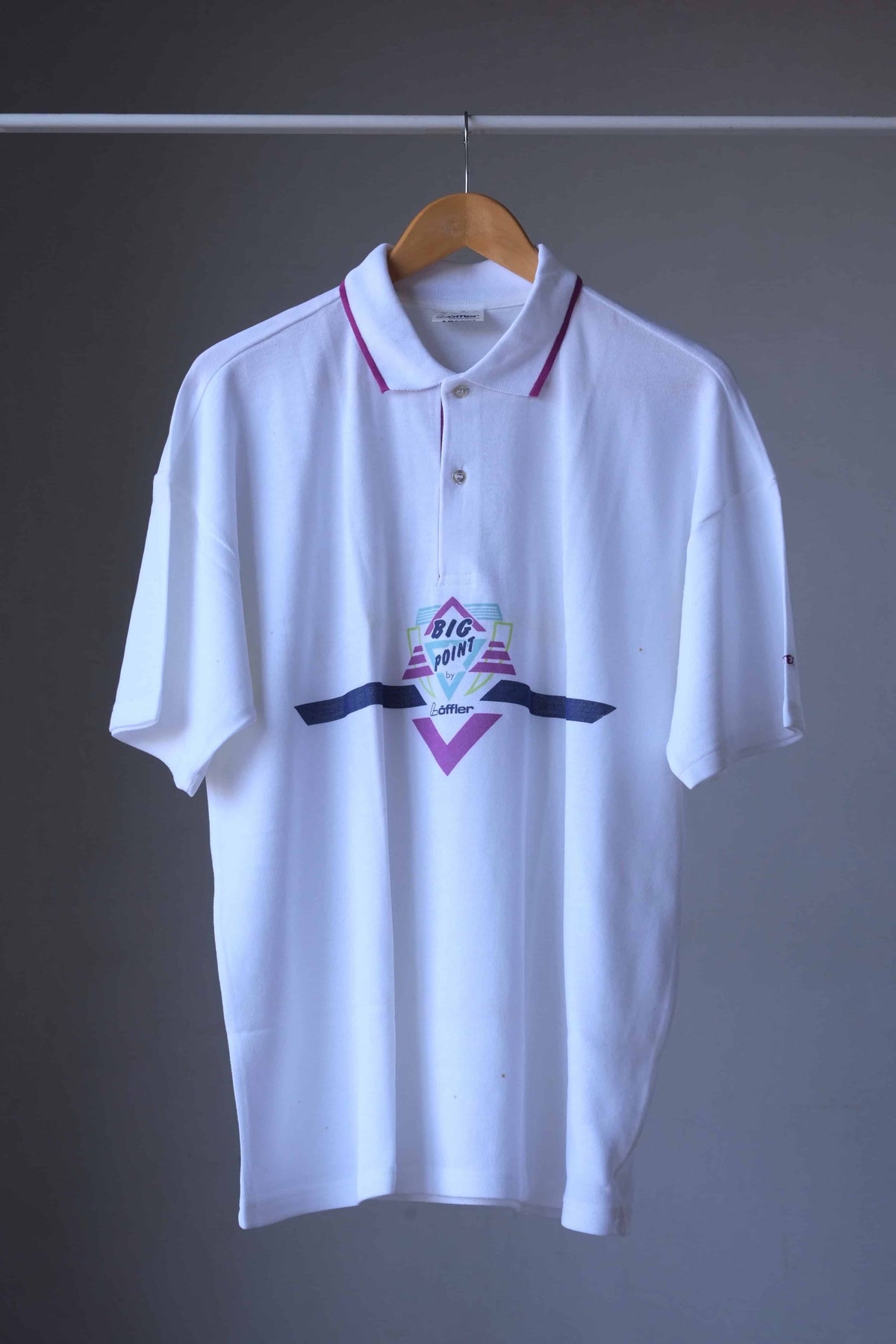 Löffler vintage 90s short-sleeve polo shirt hanging on a wooden hanger against a dark gray wall. The shirt features a small colorful graphic on the chest with a 'big point' graphic and a dark navy horizontal stripe.