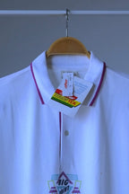 A white short-sleeve polo shirt with purple trim on the collar and placket, hanging on a wooden hanger against a gray wall. The shirt has a 'BIG POINT' logo printed on the lower front and has retail tags attached. The hang tag is visible and the words Transtex is printed on the tag.