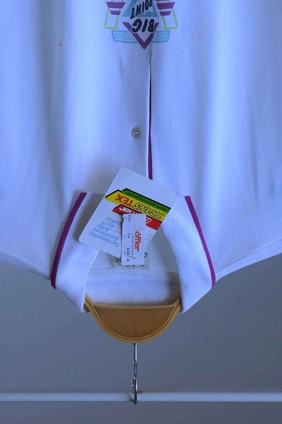 A white short-sleeve polo shirt with purple trim on the collar and placket, hanging on a wooden hanger against a gray wall. The shirt has a 'BIG POINT' logo printed on the lower front and has retail tags attached. The hang tag is visible and the words Transtex is printed on the tag.