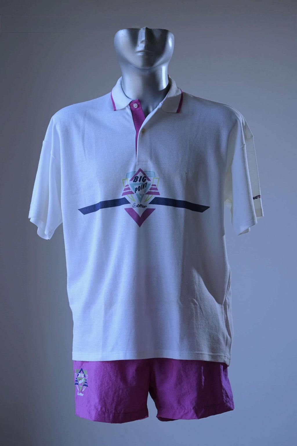 A vintage white Löffler short-sleeve polo shirt with a purple and pink collar, featuring a graphic chest design, displayed on a mannequin with matching purple shorts against a gray background.