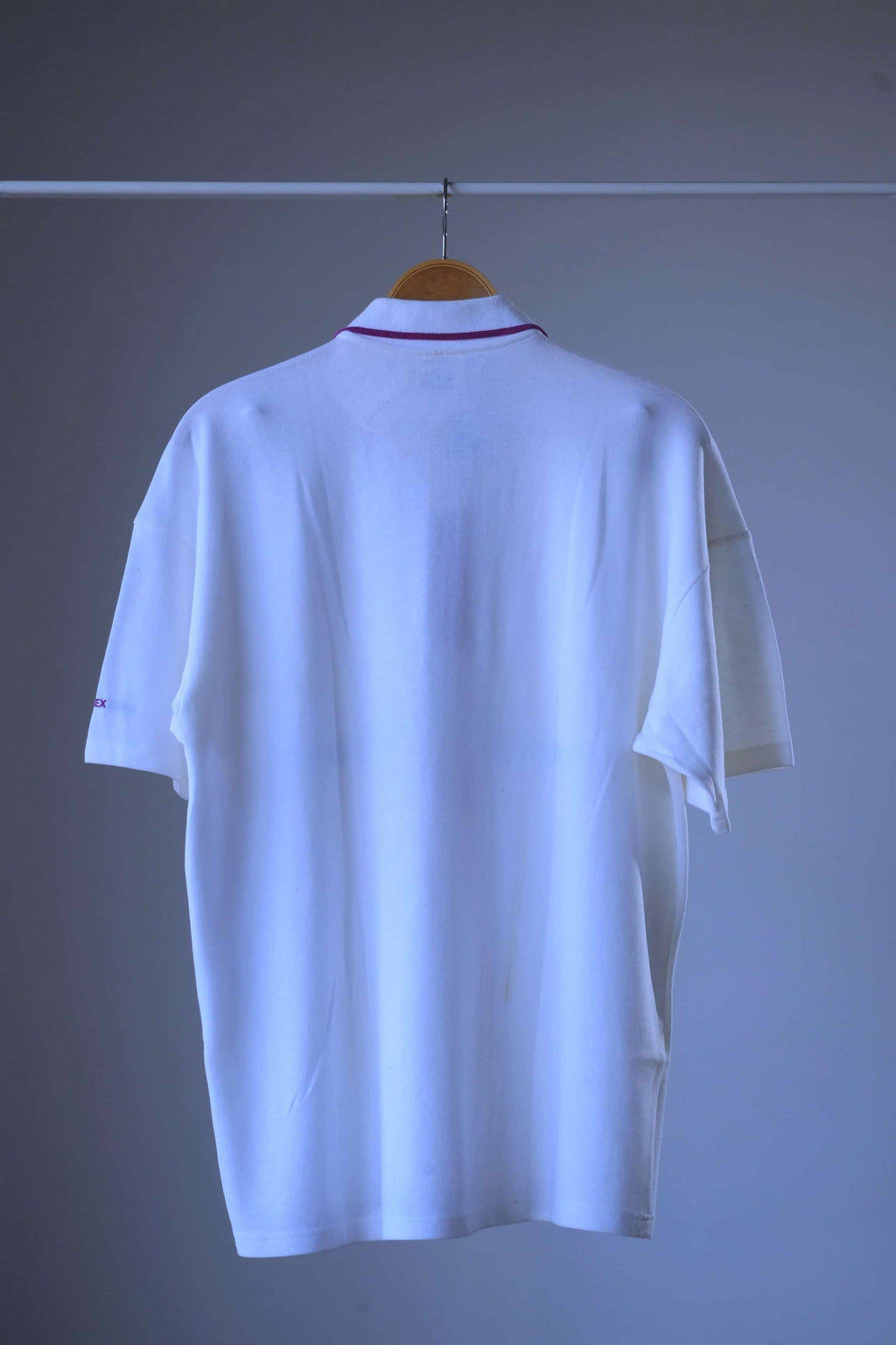 Back view of a vintage 90s Löffler white short-sleeve polo shirt hanging on a wooden hanger against a plain gray wall, showing the back view with a thin red stripe detail at the collar.