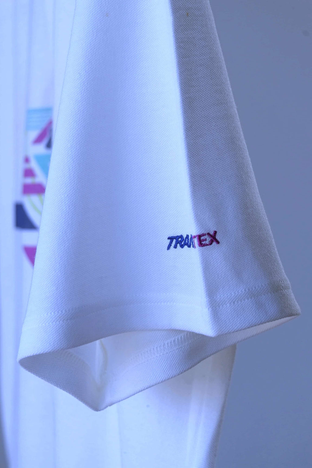 A close-up of a vintage Löffler white polo shirt sleeve featuring a small embroidered logo that reads 'Transtex' in blue and purple, set against a matching white shirt background.