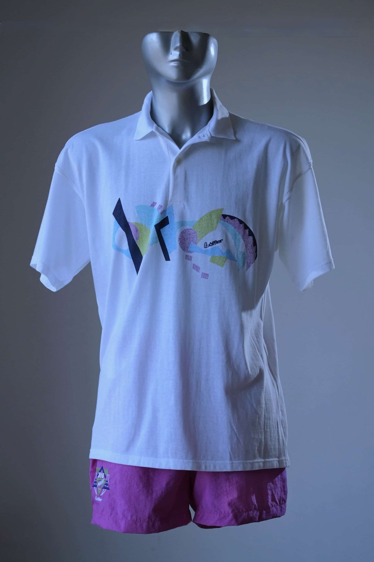 Vintage 90s Löffler men’s white tennis polo with a Memphis print in navy, sky blue, sage and lilac across the chest, displayed on a mannequin torso with purple shorts against a gray studio background.