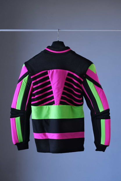 Back view of a vintage 90s Löffler slalom ski sweater hanging on a black hanger. The sweater features bold neon pink and green panels contrasted with black, padded arms and elbows, and a half-zip collar. The vibrant geometric design is prominent against the simple background.