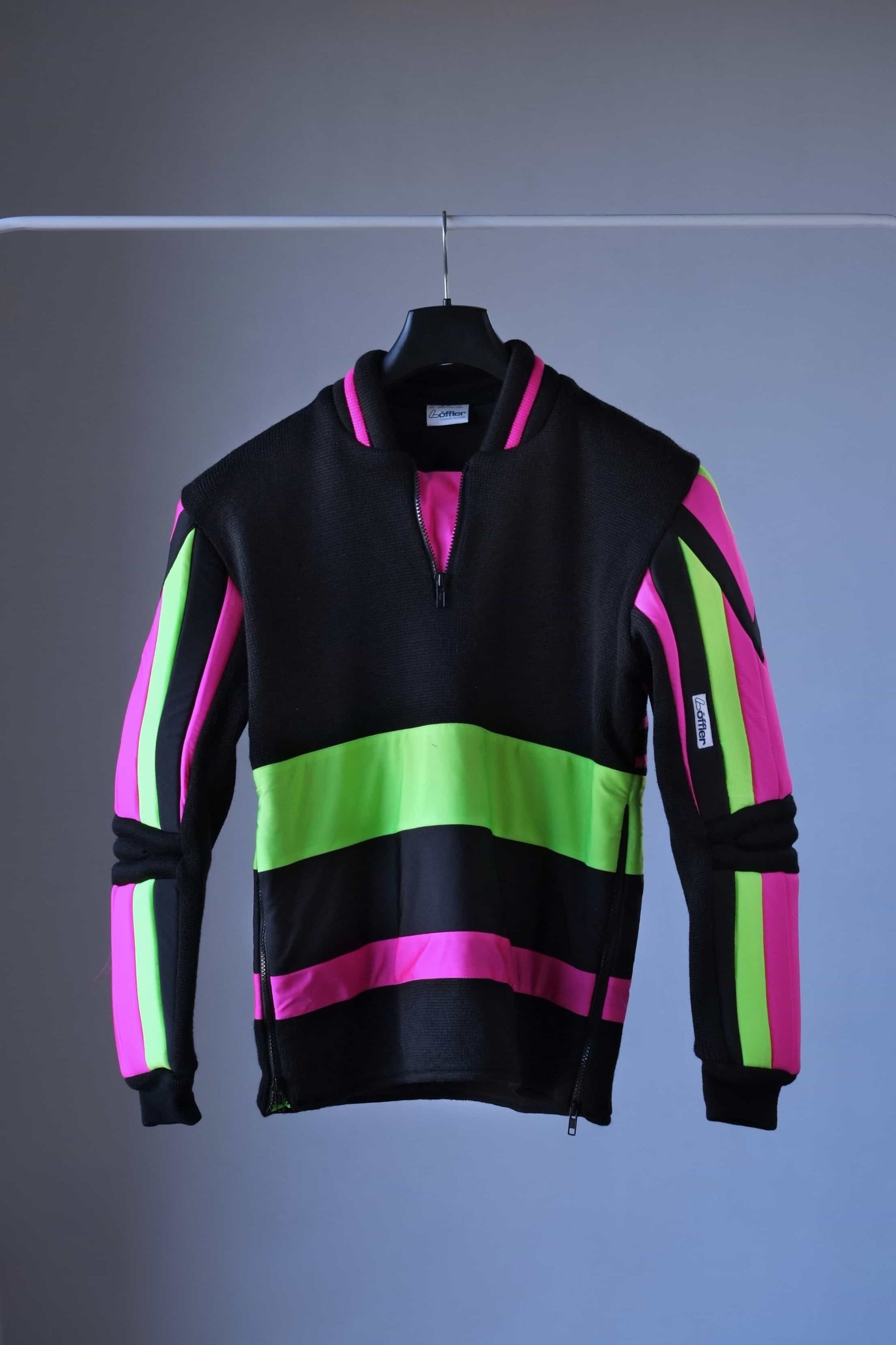 Front view of a vintage 90s Löffler slalom ski sweater hanging on a black hanger. The sweater features bold neon pink and green panels contrasted with black, padded arms and elbows, and a half-zip collar. The vibrant geometric design is prominent against the simple background.