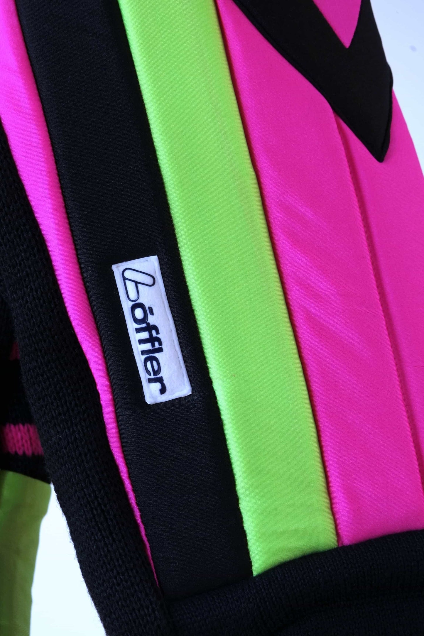 Close shot of the sleeve of a black Löffler ski sweater, featuring neon pink and green padded sections, a black zipper, and the Löffler brand label.