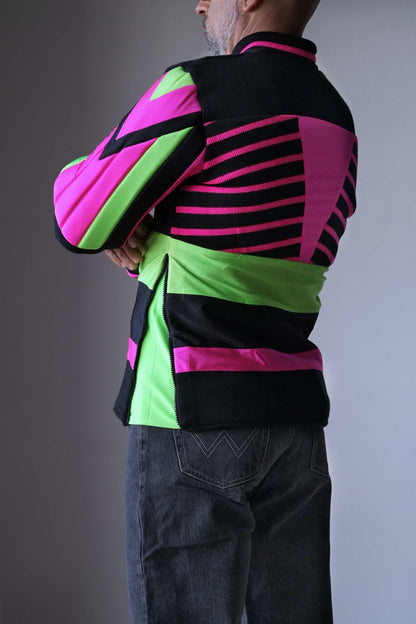 Back view of a model wearing a vintage 90s Löffler slalom ski sweater, showcasing the geometric neon pink and green design with bold black stripes on the lycra back panel. The padded arms and elbows with angular detailing are visible, along with the zipped side panel for enhanced mobility.