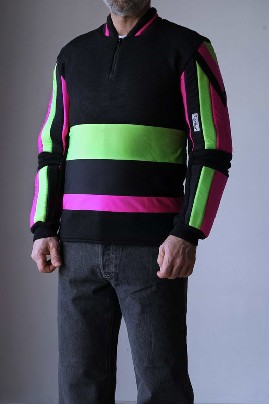 Model wearing a vintage 90s Löffler slalom ski sweater with padded arms and elbows, featuring bold neon pink and green geometric stripes on a black wool upper and lycra lower section. The sweater includes a zip neck with a cotton inset and zipped sides for added mobility and comfort.