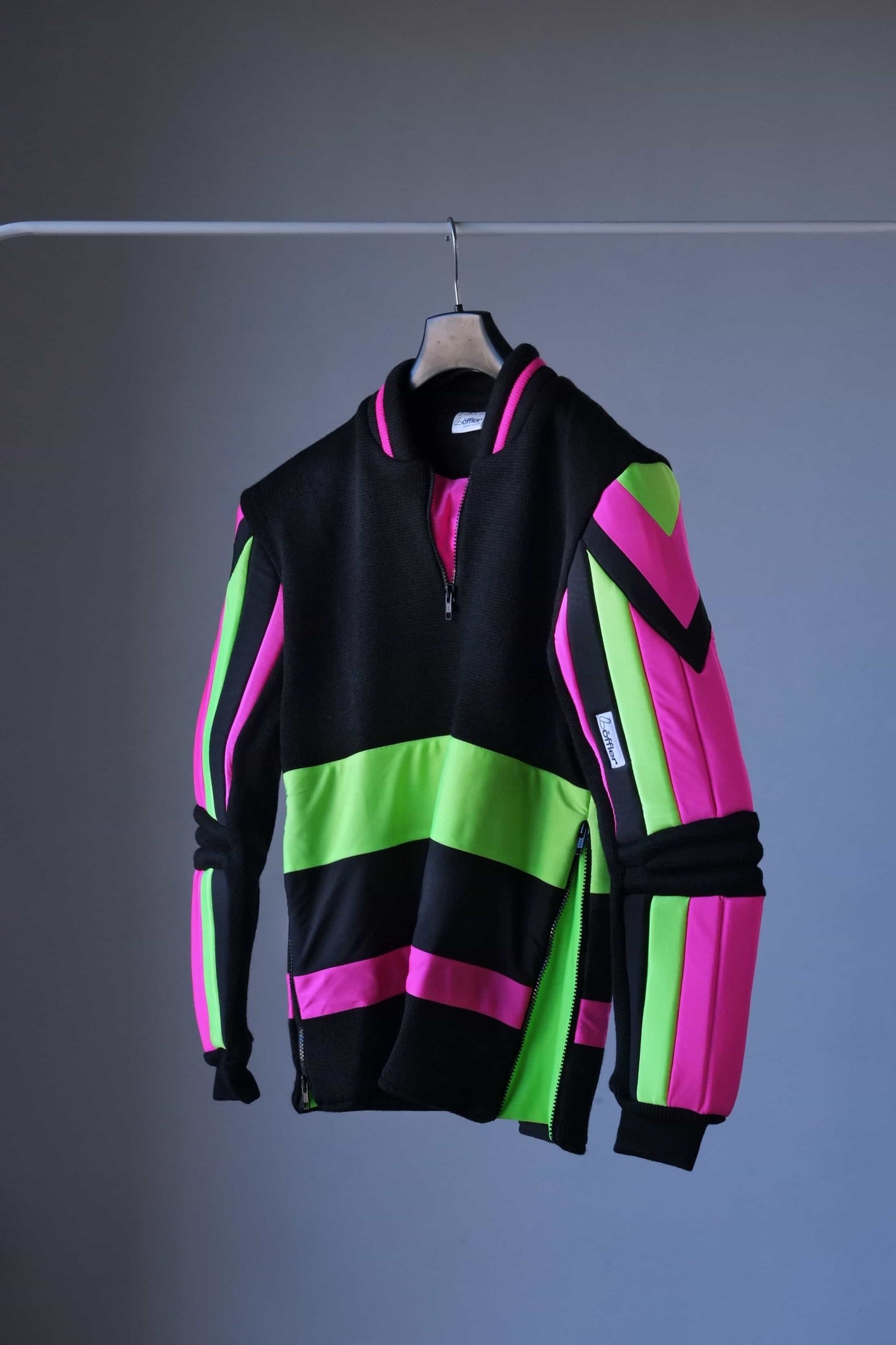 Side view of a vintage 90s Löffler slalom ski sweater hanging on a black hanger. The sweater features bold neon pink and green panels contrasted with black, padded arms and elbows, and a half-zip collar. The vibrant geometric design is prominent against the simple background.