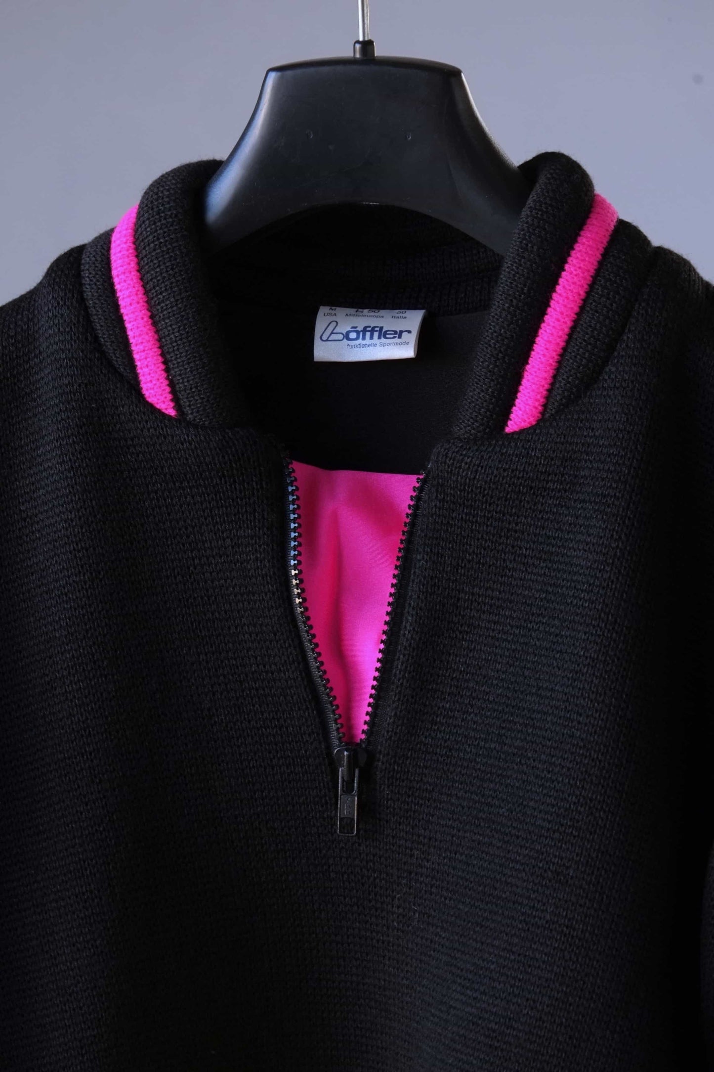 Close-up of the front of a black Löffler ski sweater, featuring a padded collar with pink accents and inset, and a black zipper. The label can be seen with the word Löffler on it.