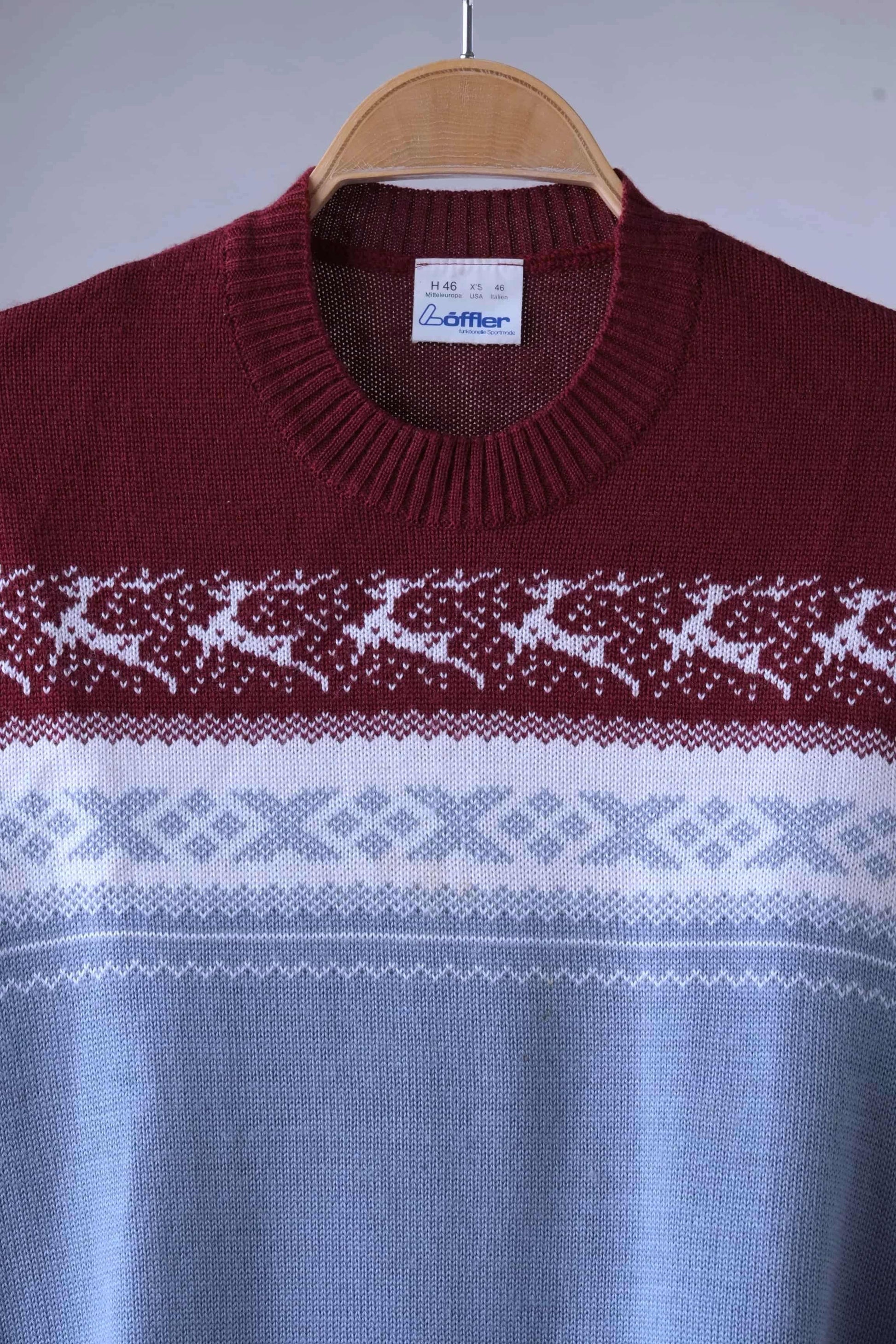 Close view of Löffler brand burgundy and gray wool blend sweater featuring a white Nordic pattern with reindeer and snowflake motifs, hanging on a wooden hanger against a plain background.