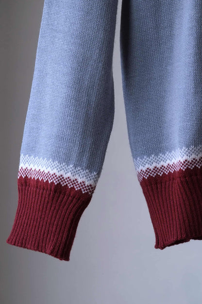 Close up of the sleeve of a Löffler brand burgundy and gray wool blend sweater featuring a white Nordic pattern with reindeer and snowflake motifs, hanging on a wooden hanger against a plain background.
