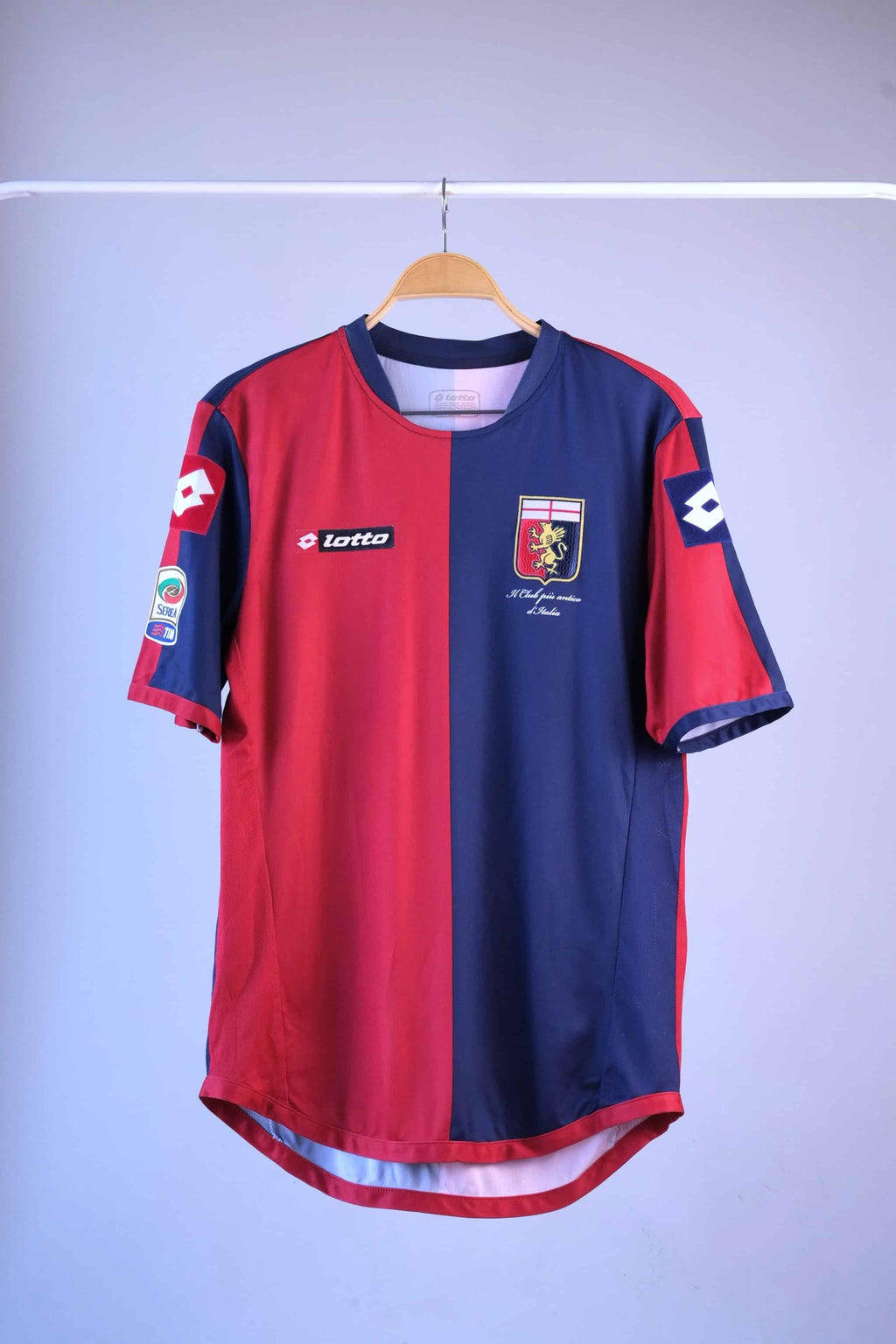 Front view of a red and navy blue vertically split football jersey hanging on a wooden hanger against a plain grey background. The jersey features a black "lotto" logo on the right breast, a club crest on the left breast, and a Serie A TIM patch on the right sleeve.