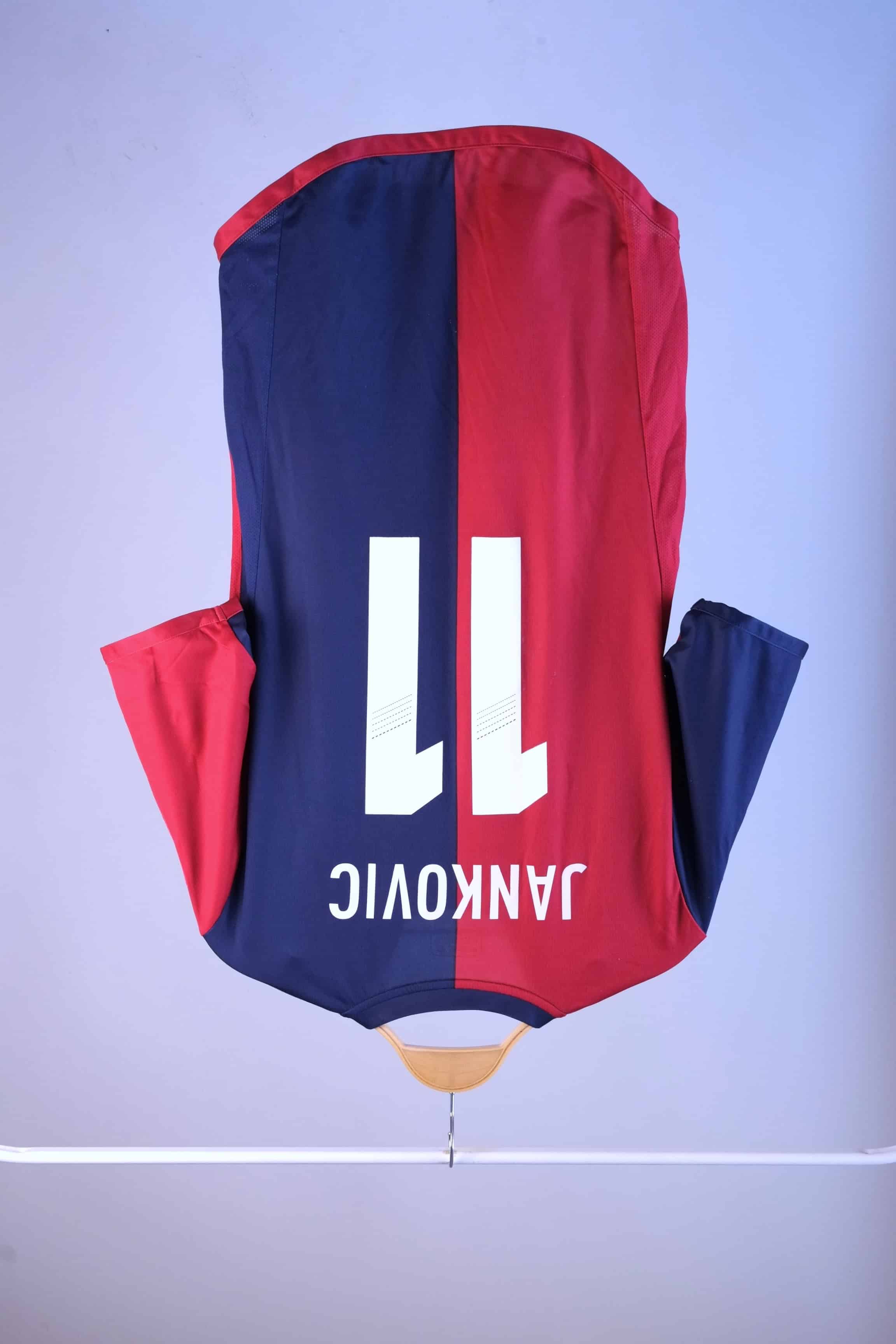 Back view of a red and navy blue vertically split football jersey hanging on a wooden hanger against a plain grey background. The name "JANKOVIC" and the number "11" are printed in large white text across the upper back.
